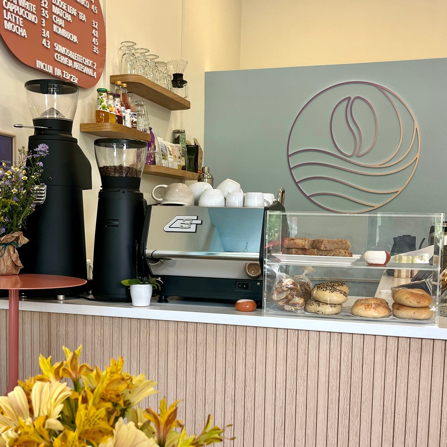 The cozy cafeAtaraxia interior featuring a wooden counter with a coffee machine, espresso dispensers, and a glass display case filled with bagels and pastries in Parede